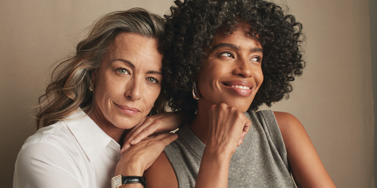 Lighter skin tone model faces the camera and leans on a dark skin model who looks outward