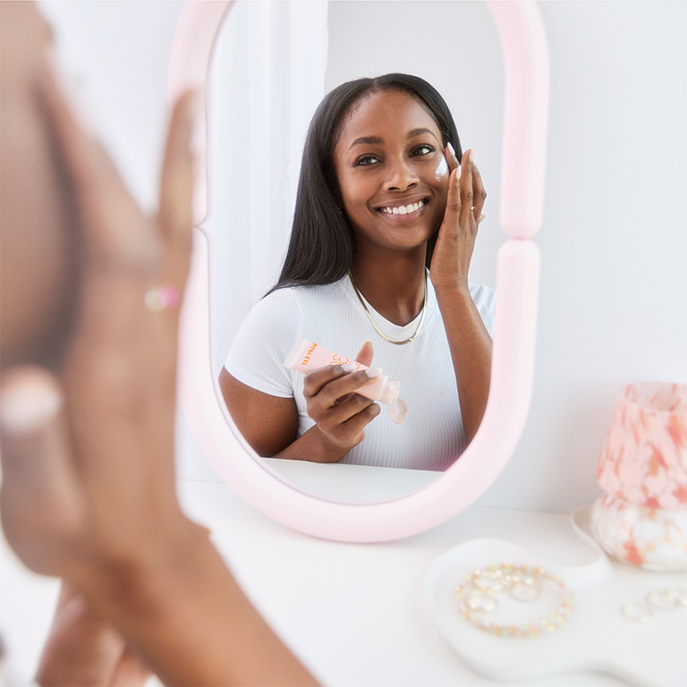 Woman looking into a makeup mirror and apply Mary Kay Mineral Facial Sunscreen Broad Spectrum SPF 30 to her cheek